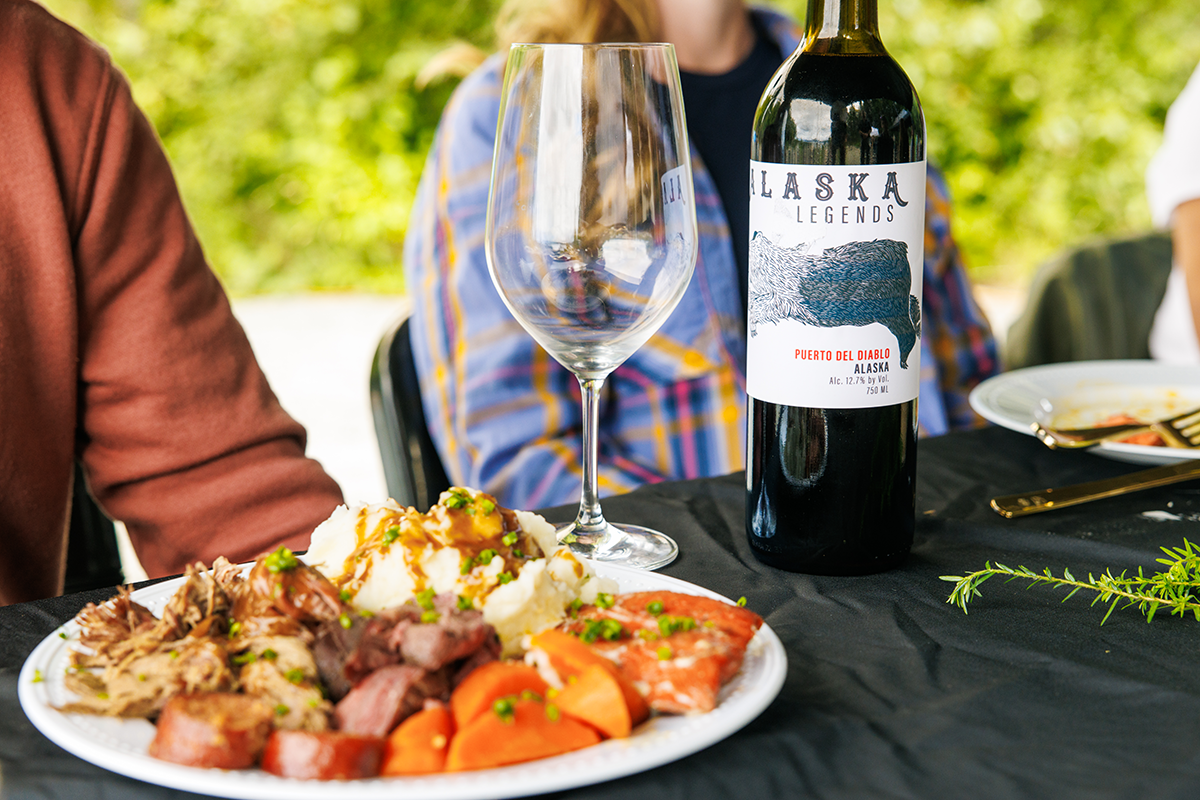 Outdoor table with a plate of roast meat, mashed potatoes and vegetables, a bottle of Alaska Legends wine, and an empty wine glass nearby.