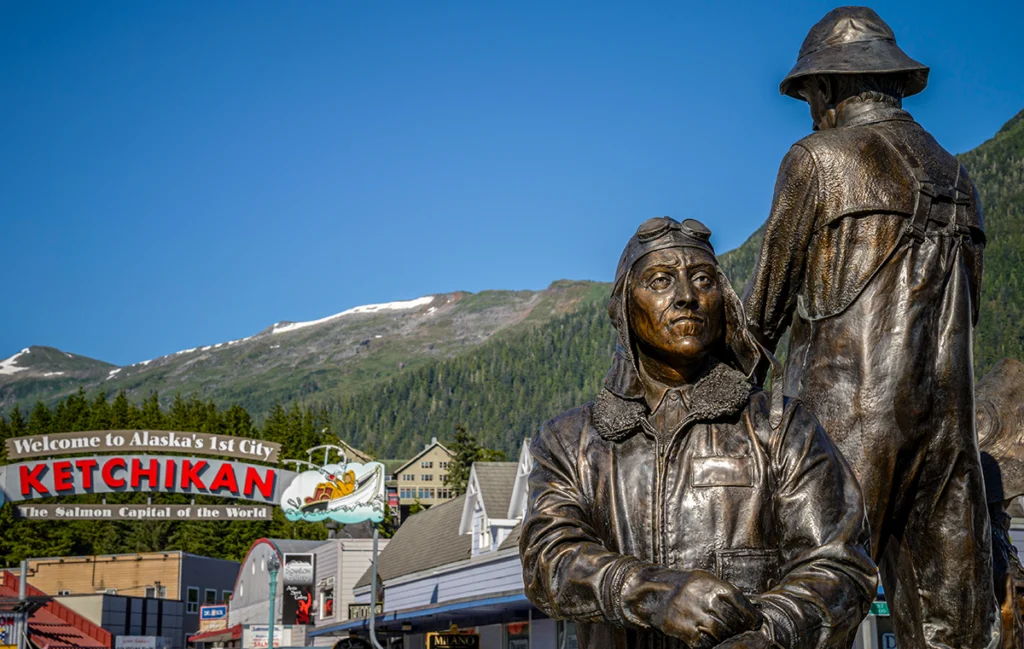 Bronze statues stand in front of a “Welcome to Alaska’s 1st City Ketchikan” sign with mountains behind them.