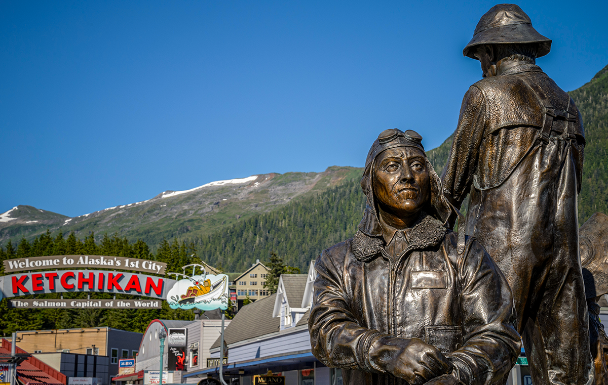 Bronze statues stand in front of a “Welcome to Alaska’s 1st City Ketchikan” sign with mountains behind them.