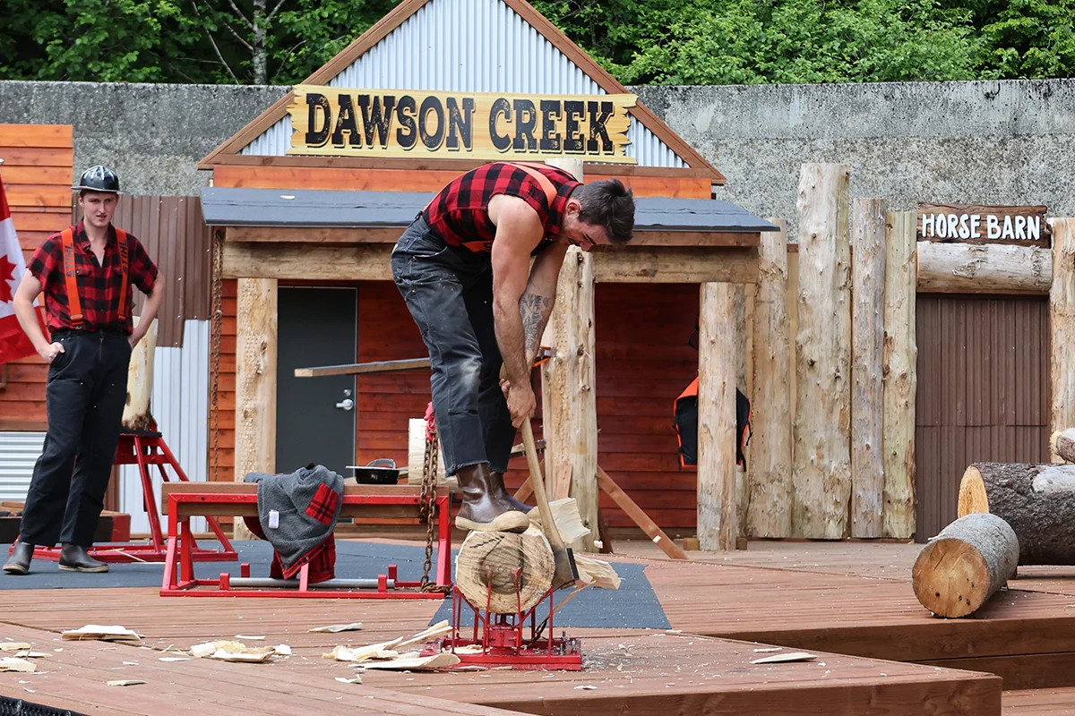 Man in a red plaid shirt splitting a log with an axe on a wooden platform, while another man watches in the background; Dawson Creek sign visible.