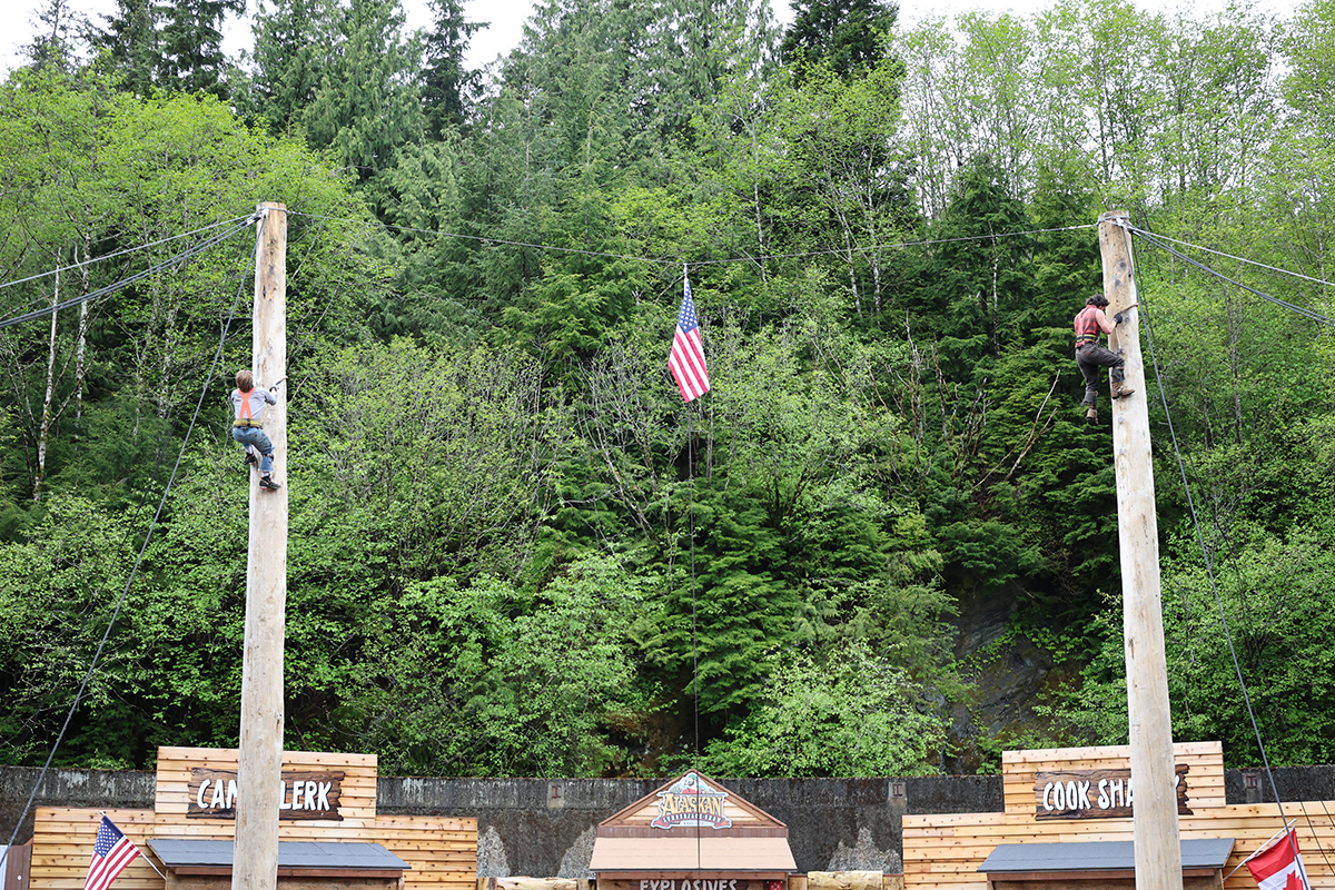Two climbers in harnesses scale tall wooden poles on a high-ropes course, with an American flag between them and green forest in the background.