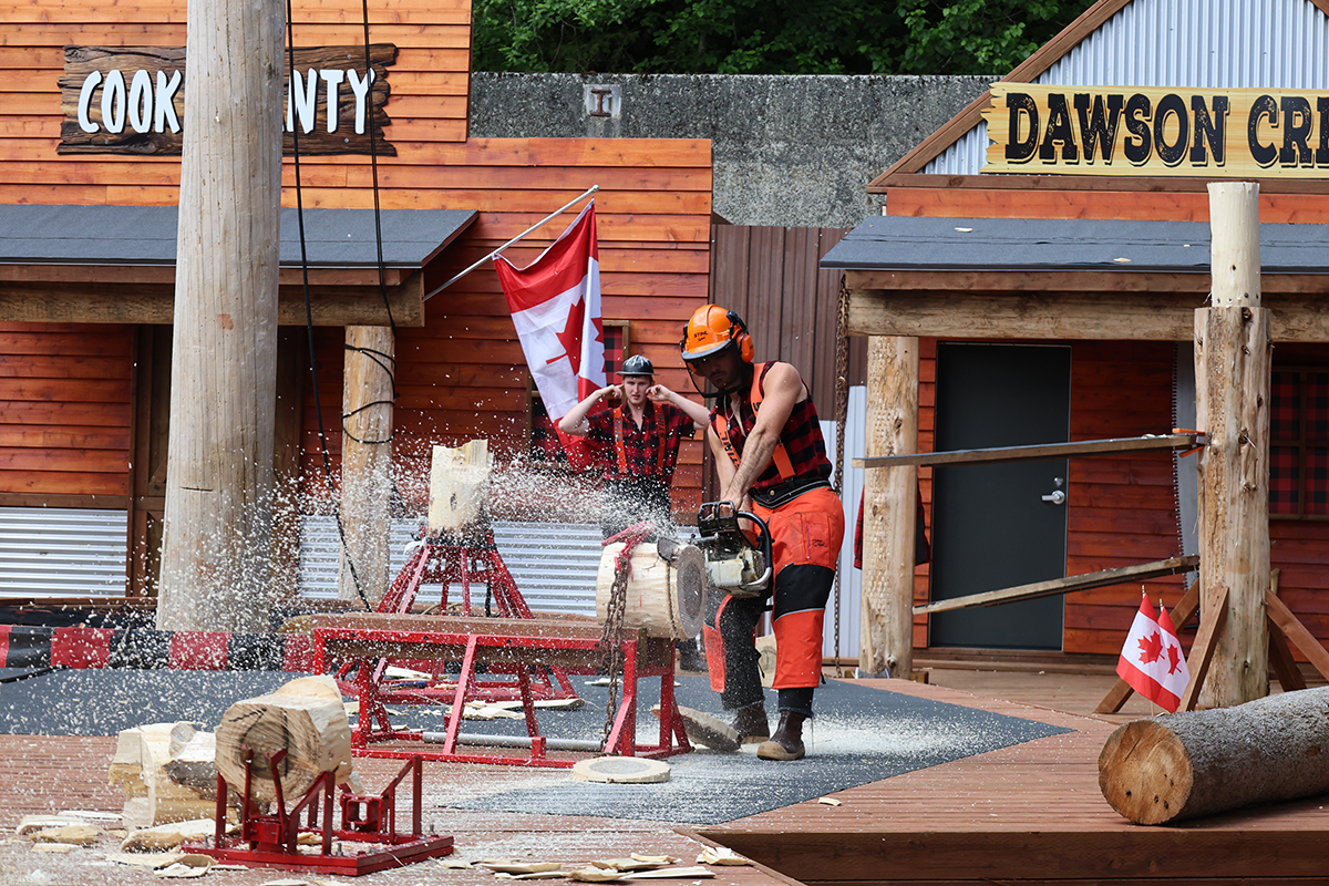 Two lumber workers in orange and red safety gear cut a log with a chainsaw as sawdust flies, outdoors beside wooden buildings with Canadian flags nearby.