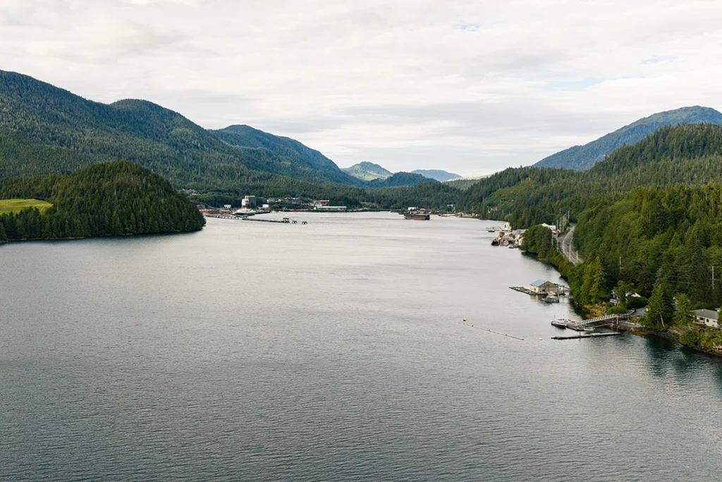 A scenic harbor with mountains, forested shoreline, and industrial port buildings along the water.