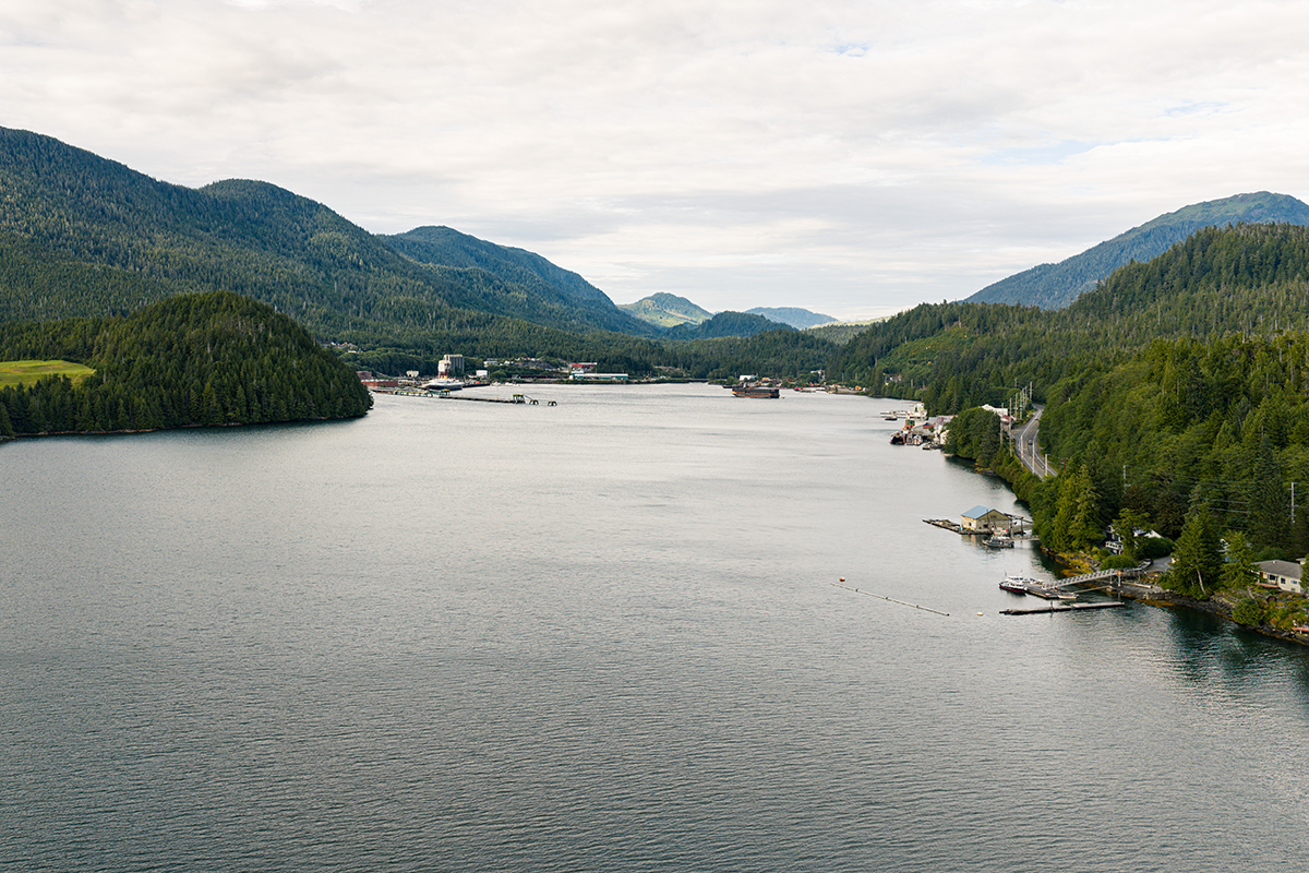 A scenic harbor with mountains, forested shoreline, and industrial port buildings along the water.